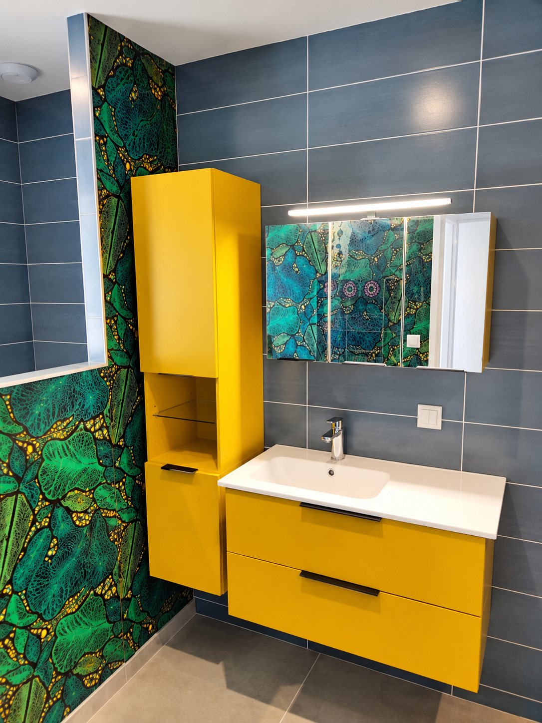 Meuble de salle de bain jaune et colonne assortie devant un mur à motif feuillage